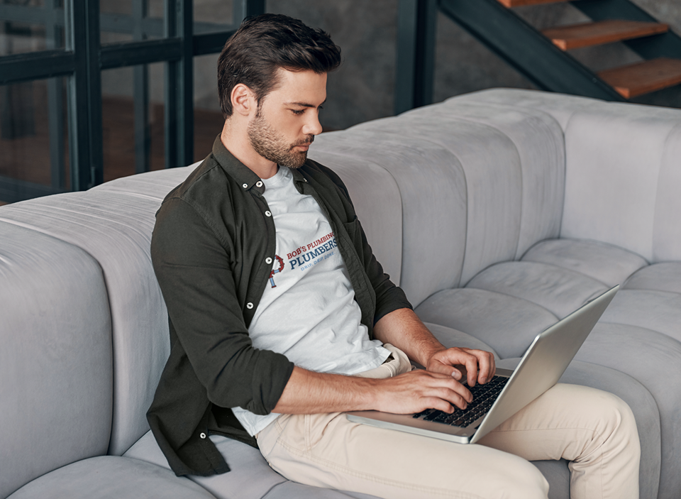 A man in his late 20s wearing a white ‘Bob’s Plumbing’ T-shirt and dark open shirt, sitting on a light grey couch and typing on a laptop, illustrating the concept of does online marketing work for small businesses.