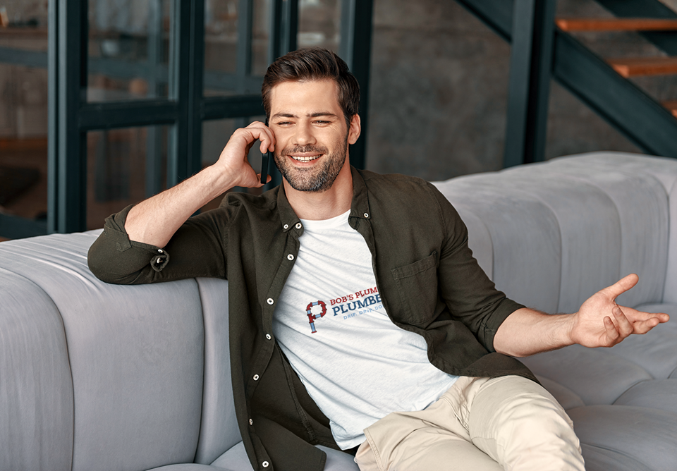 A man in his late 20s wearing a white ‘Bob’s Plumbing’ T-shirt and dark open shirt, sitting on a light grey couch while talking on a mobile phone, illustrating the concept of does online marketing work for small businesses.